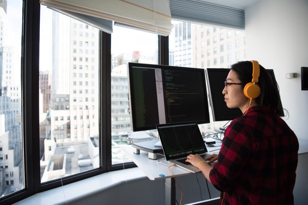 Woman sitting at her laptop and desktop, appearing to write code. She is wearing bright yellow headpones.