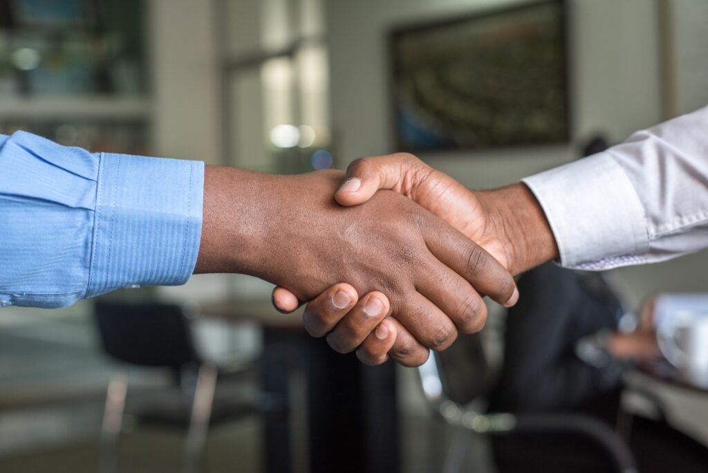 Two people shaking hands, one wearing a blue long-sleeve dress shirt, the other wearing a white long-sleeve dress shirt.