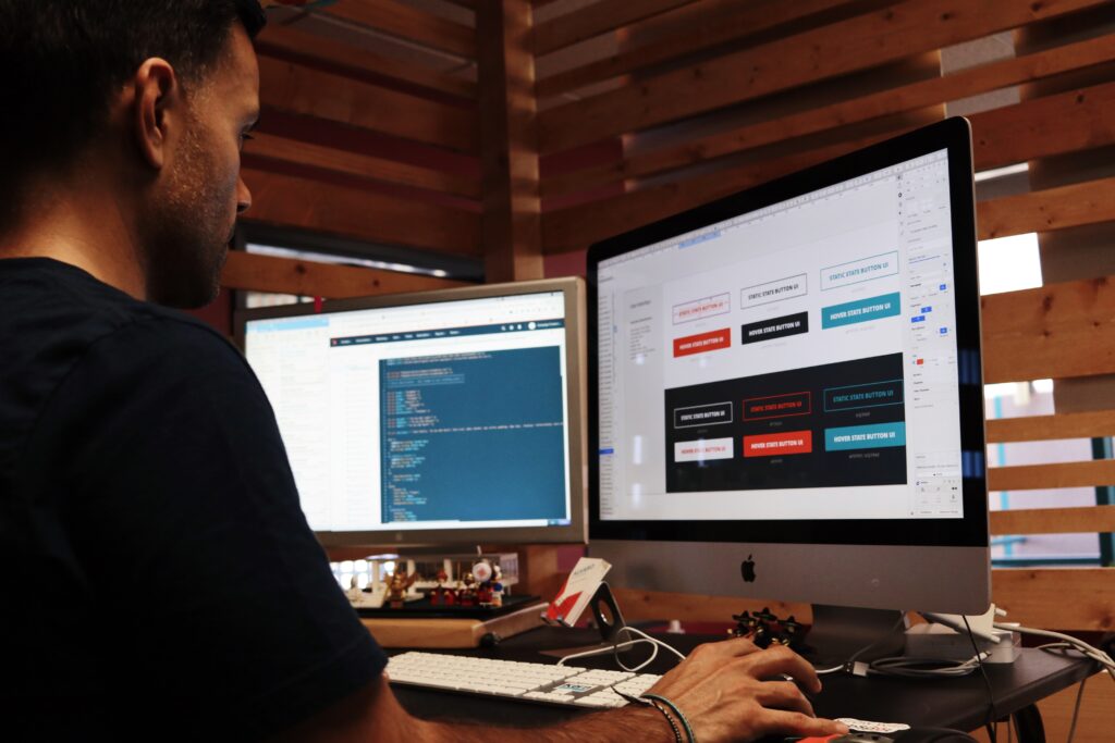 A man sitting down at a desk behind two monitors working on them. It looks like he is programming a website.