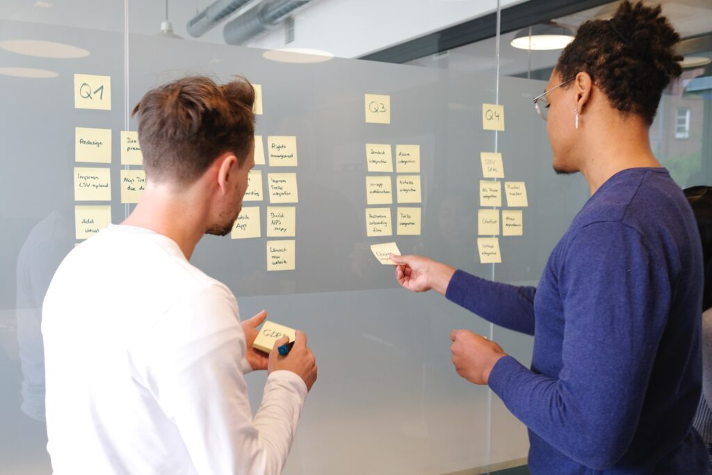 Man in blue long sleeve shirt standing in front of a wall with another man in a white shirt. They are looking at post-it notes on a wall that seem to have to do with different quarters of the business they work for.