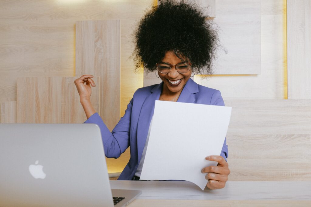 A woman with a purple blazer sitting behind her laptop looking at a stack of papers with a big smile on her face.