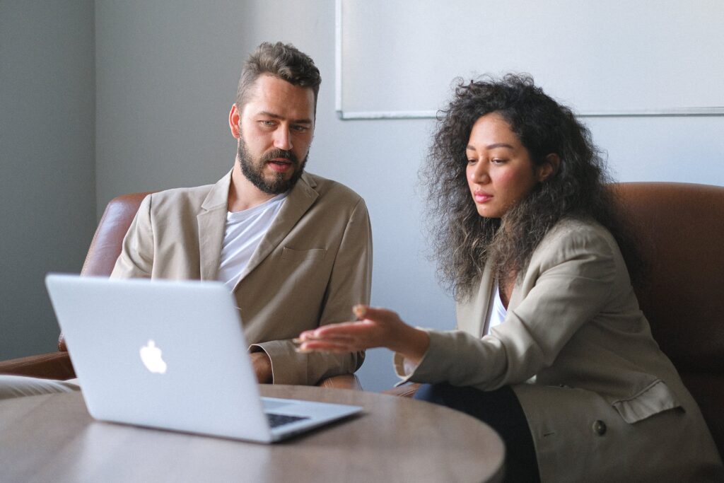 A man and a woman in blazers sitting behind a laptop screen that they are both looking at. It seems as though they are having a work-related meeting.