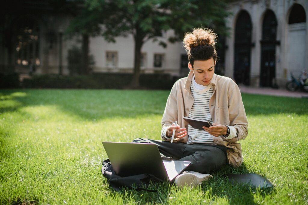 A man with tied up hair sitting on a lawn working at his computer in university.
