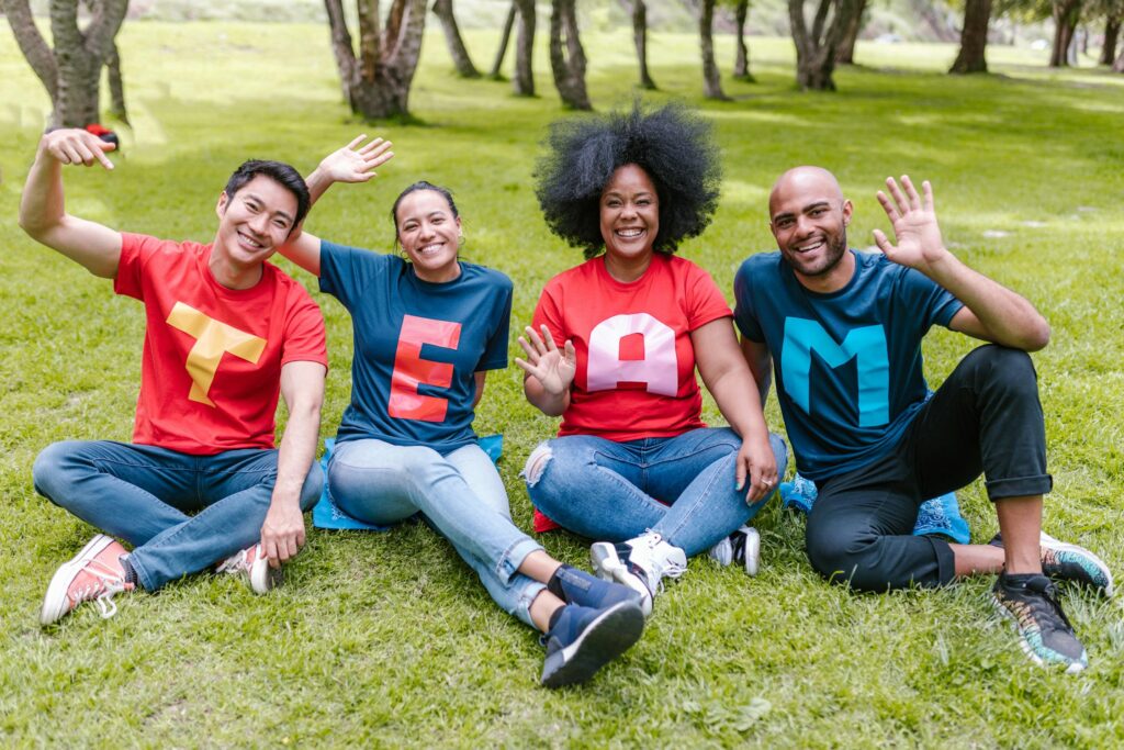 Four people sitting on a lawn smiling, each wearing a shirt with a letter on it, together spelling "TEAM"