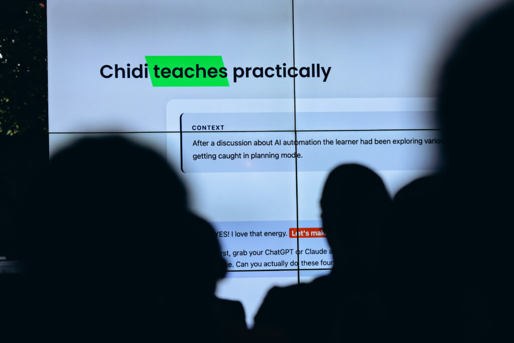 The shadows of attendee's heads in the front of the frame, they are all facing a big screen that says "Chidi teaches practicality" during a presentation at the ALX x Anthropic event in Kigali, Rwanda. 